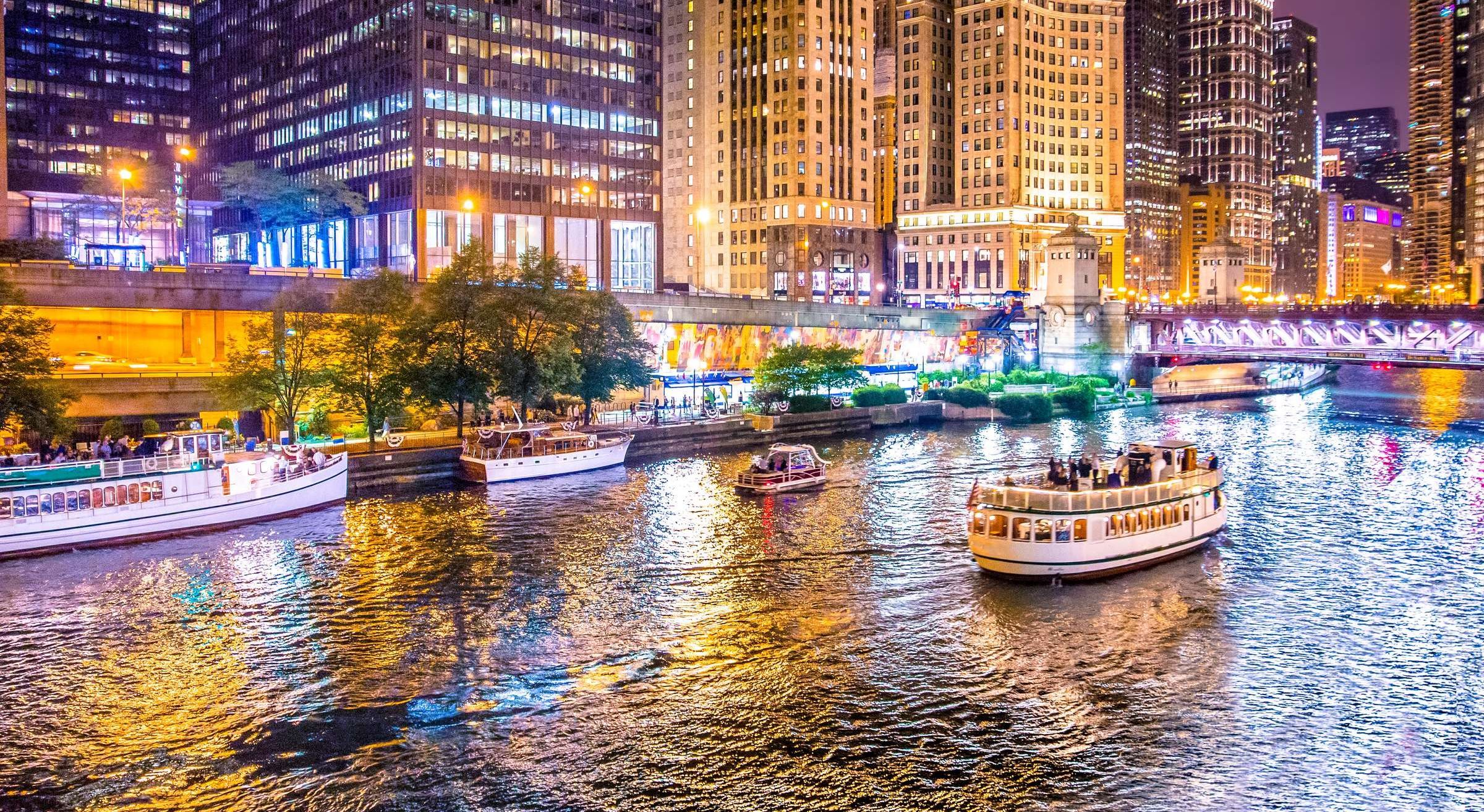 Optima Signature in Downtown Chicago with Tour boats on a brightly lit city river at night, surrounded by tall buildings and reflections on the water.
