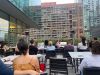 Optima Signature in Downtown Chicago with People at an Optima Communities rooftop patio enjoy a celebration as a woman speaks on a microphone with city buildings behind her.