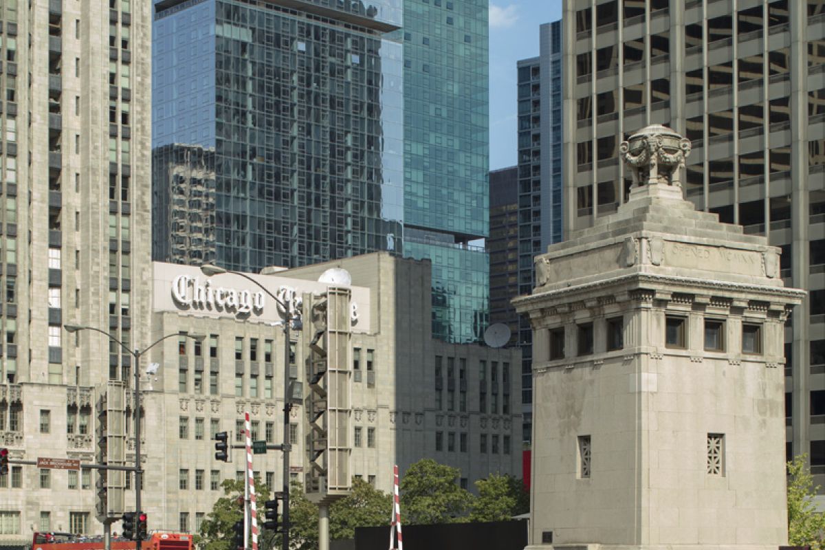 Optima Signature in Downtown Chicago with A man stands on a city bridge, reflecting on the value of commute, with tall skyscrapers and the Chicago Tribune building behind him.