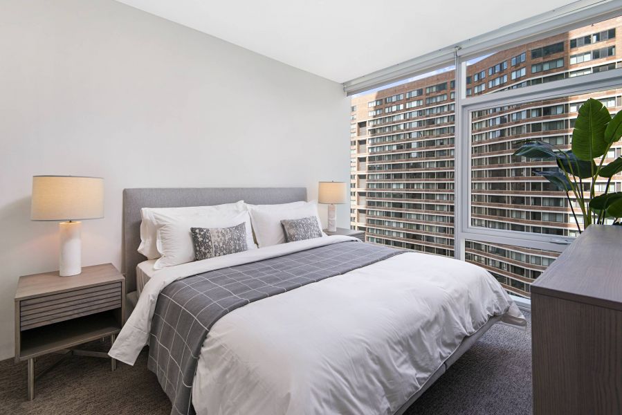 Optima Signature in Downtown Chicago with Modern bedroom with a neatly made bed, two lamps, and large windows overlooking city buildings.