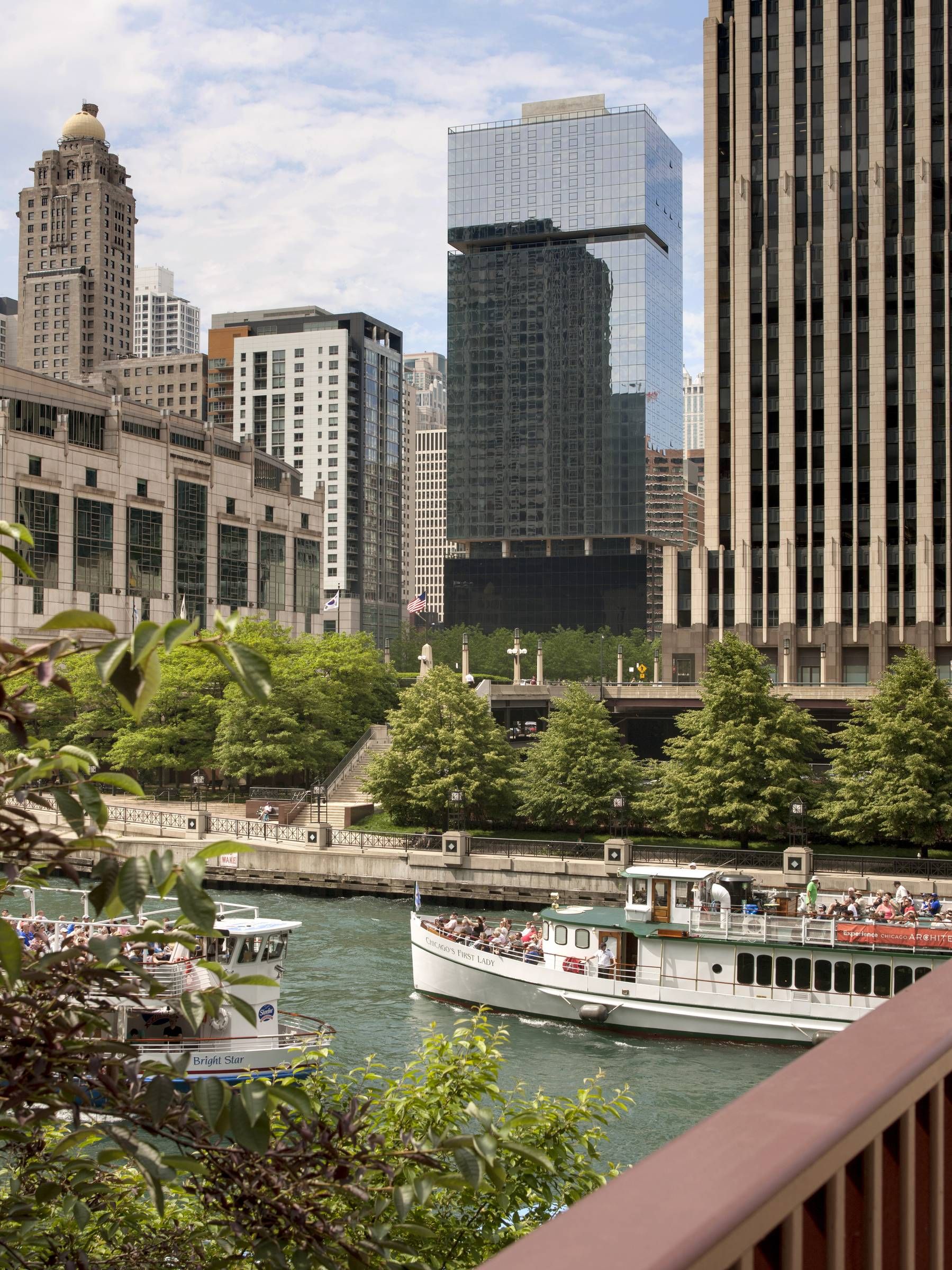 Optima Signature in Downtown Chicago with Tour boats on a river surrounded by tall city buildings and greenery on a partly cloudy day.