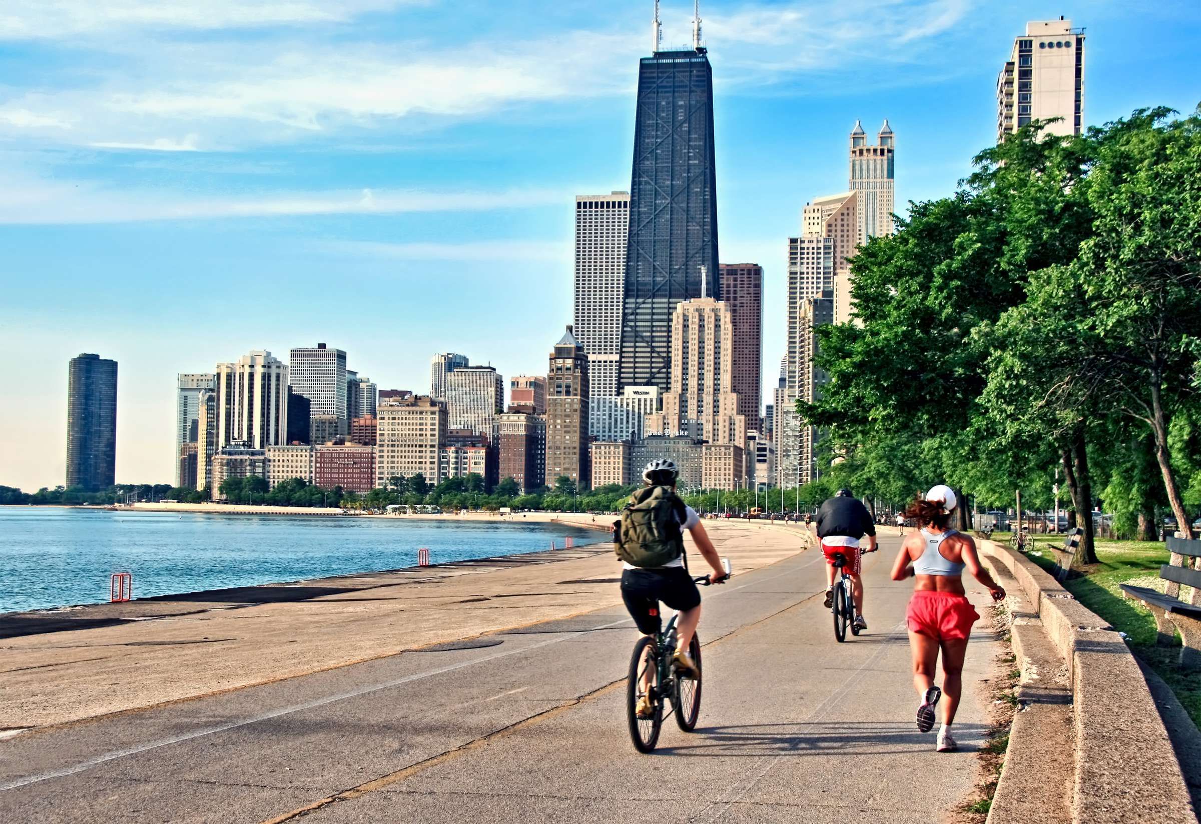 Optima Signature in Downtown Chicago with People biking and jogging on a lakeside path with Chicago skyline and trees in the background.