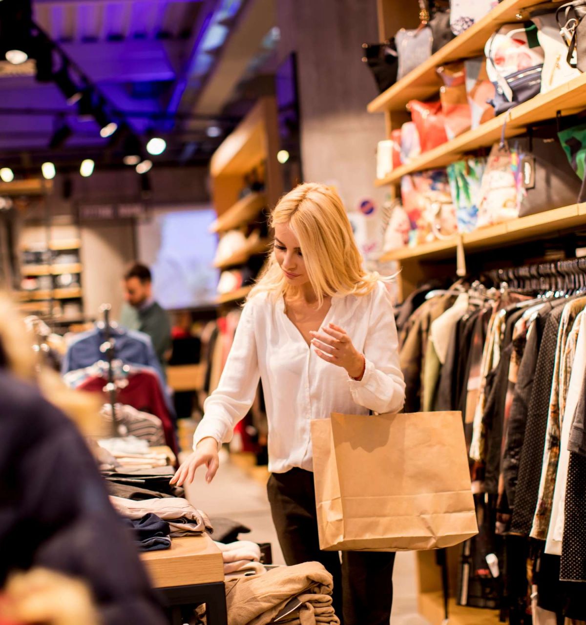 Optima Signature in Downtown Chicago with Woman shopping for clothes in a store, holding a paper bag and browsing folded shirts on a table.