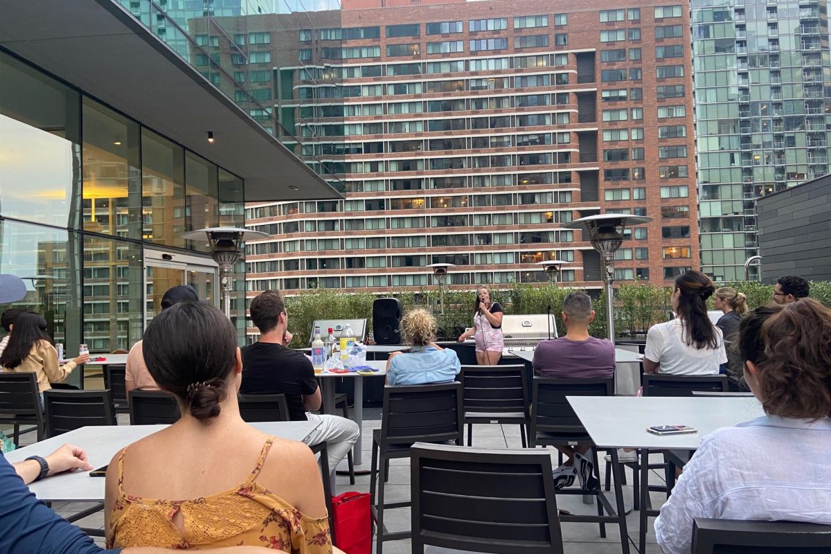 Optima Signature in Downtown Chicago with People at an Optima Communities rooftop patio enjoy a celebration as a woman speaks on a microphone with city buildings behind her.